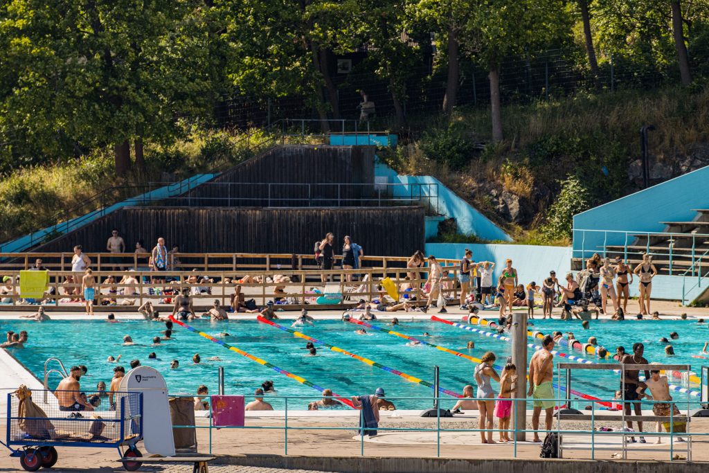 Busy outdoor public swimming pool with lane ropes, poolside seating and trees in the background.