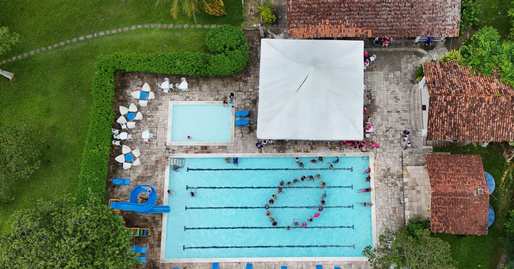 Outdoor resort swimming pool with trees, sun loungers and buildings in the background.